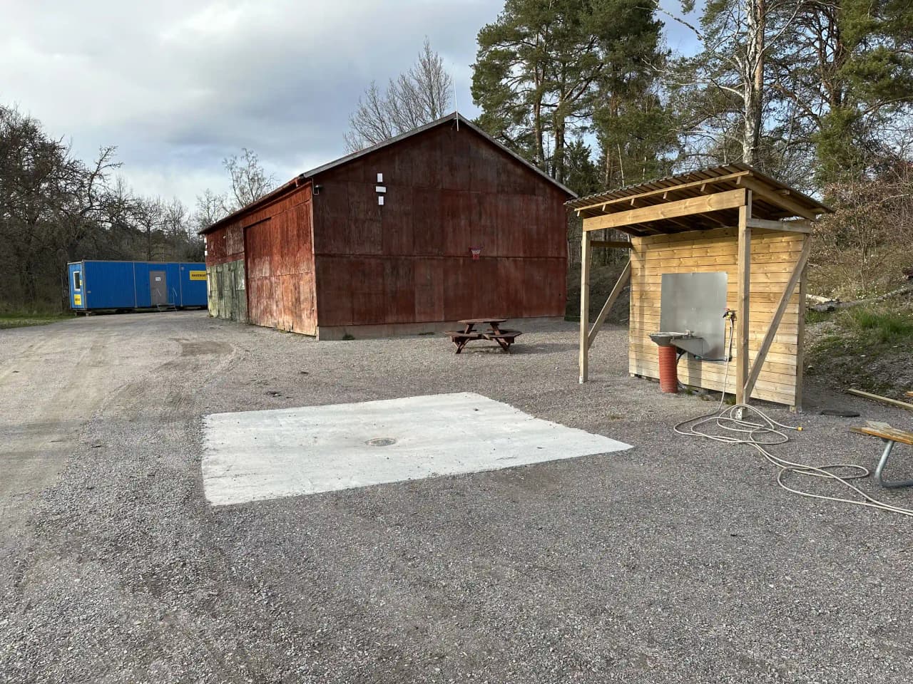 En asfalterad uppfart vid Björknäs camping omgiven av trädgårdsbyggnader och grönska, med en garagebyggnad synlig i bakgrunden.