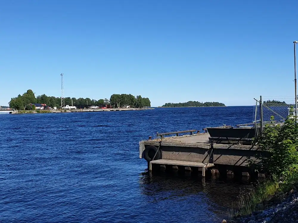En bild av Ljusnefors camping visar en naturskön plats vid vattnet, med inslag av sjölandskap och kustlinje. I bakgrunden syns en vattenväg som slingrar sig genom området, omgiven av frodig grönska och en brygga vid kanten.