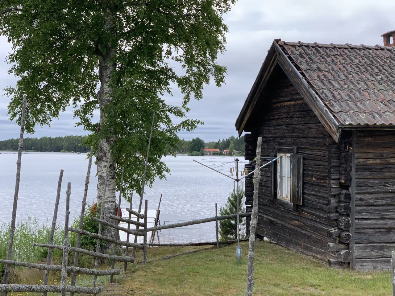 En idyllisk utsikt över Högbacka camping med en timmerstuga vid en lugn sjö, omgiven av trädkantade strandkanter och enkla trädgårdsbyggnader.