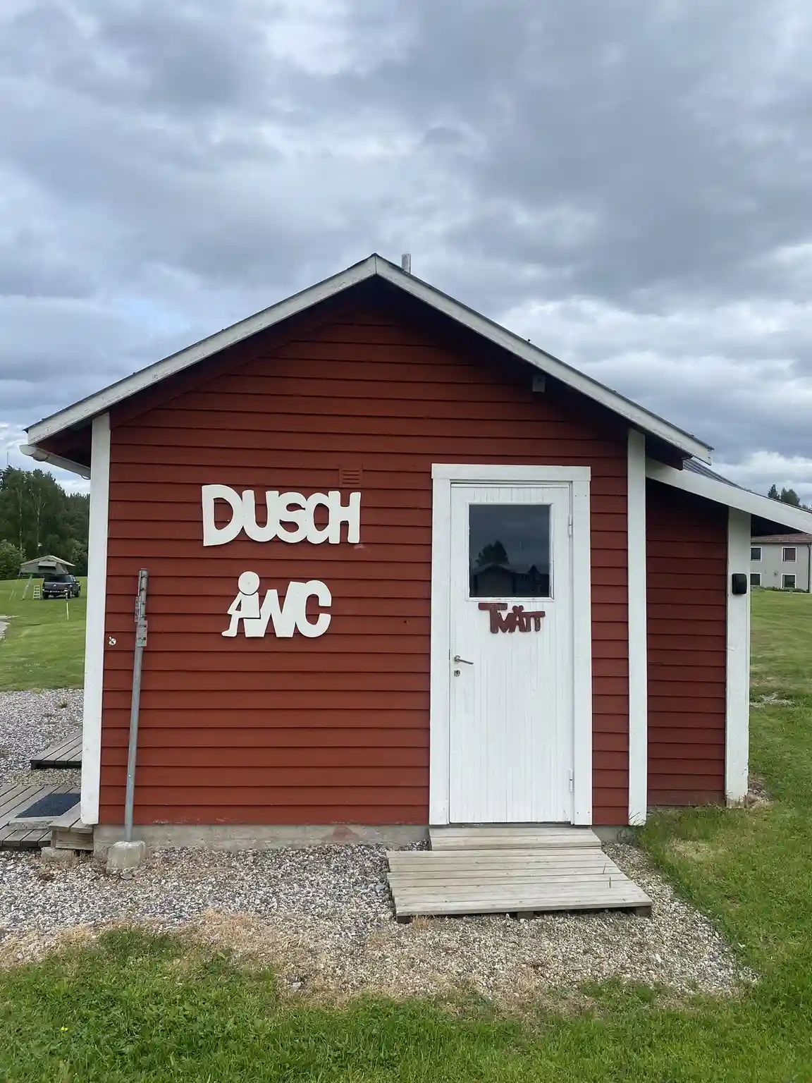 En träbyggnad vid Åmsele camping med en grön dörr och träpanel. På väggen finns en skylt. Omgivningen består av en välskött gräsyta med trädgårdsbyggnader i bakgrunden.