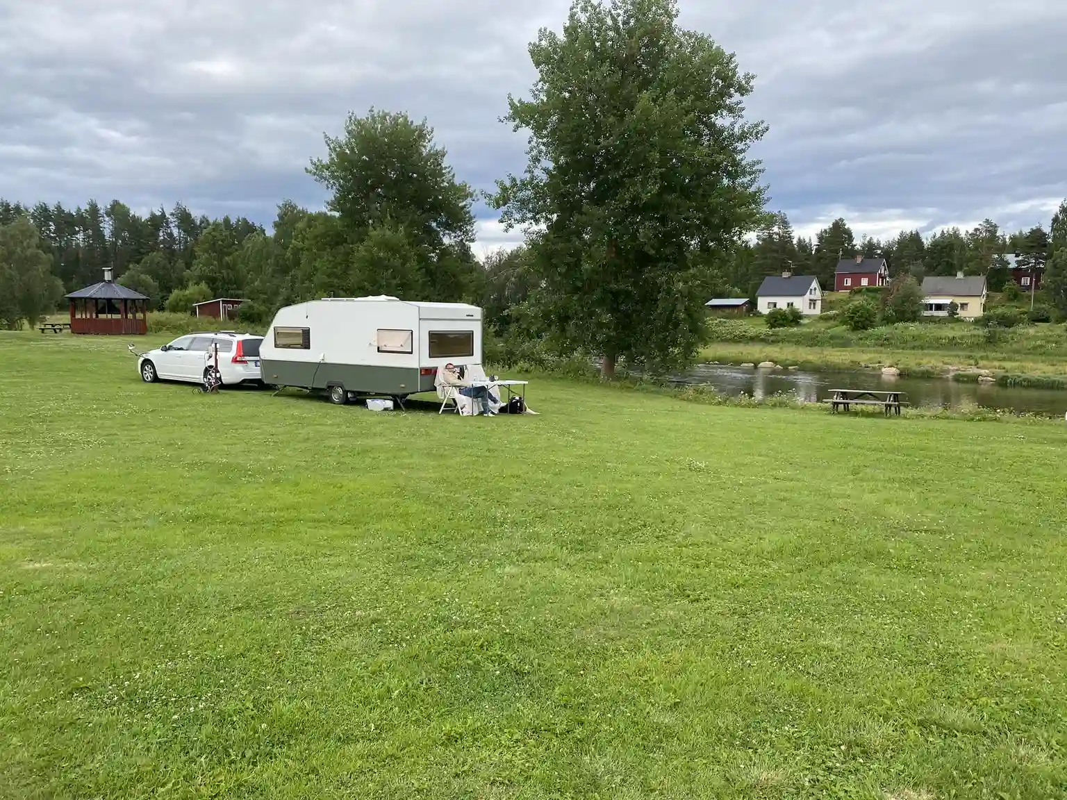 Bild på Åmsele camping med husvagnar uppställda på en gräsmatta. I bakgrunden syns ett vidsträckt område med gröna ängar och växtlighet under en delvis molnig himmel.
