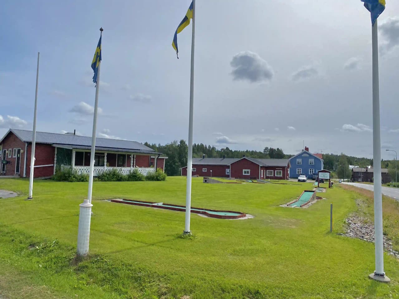 En välskött gräsyta vid Åmsele camping med en flaggstång i mitten. I bakgrunden syns en klar himmel med cumulusmoln.