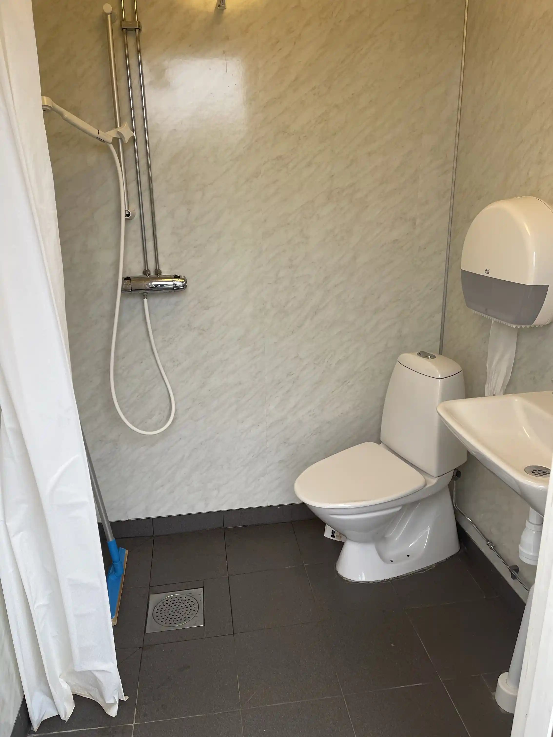 En ren och välskött toalett med vita väggar, kaklat golv och moderna sanitetsartiklar, inklusive en toalettstol och ett handfat.