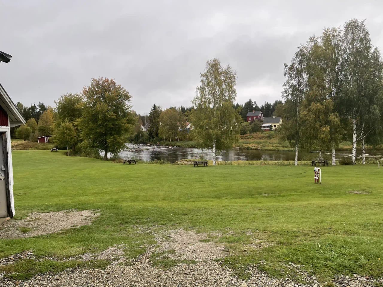 En grönskande campingplats i Åmsele med träd och gräsytor omgivna av molntäckt himmel. En damm eller liknande vattendrag syns i närheten.