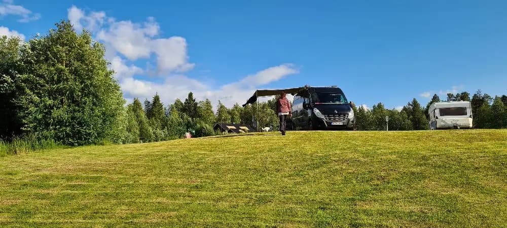 En vidsträckt grön äng med en husbil parkerad i bakgrunden under en himmel med vita cumulusmoln.