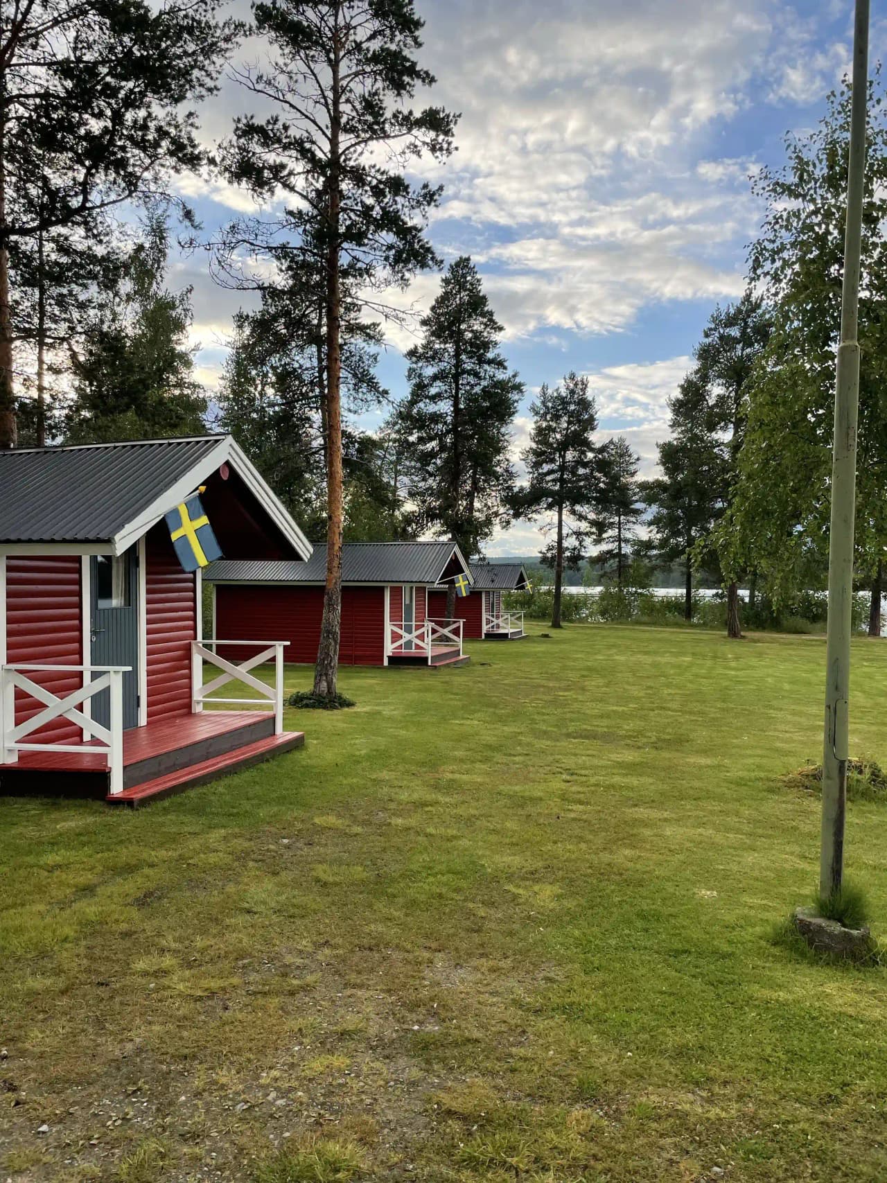 En grön gräsyta med omgivande träd, enkel campingstruktur och små byggnader i bakgrunden på Fredrika camping.
