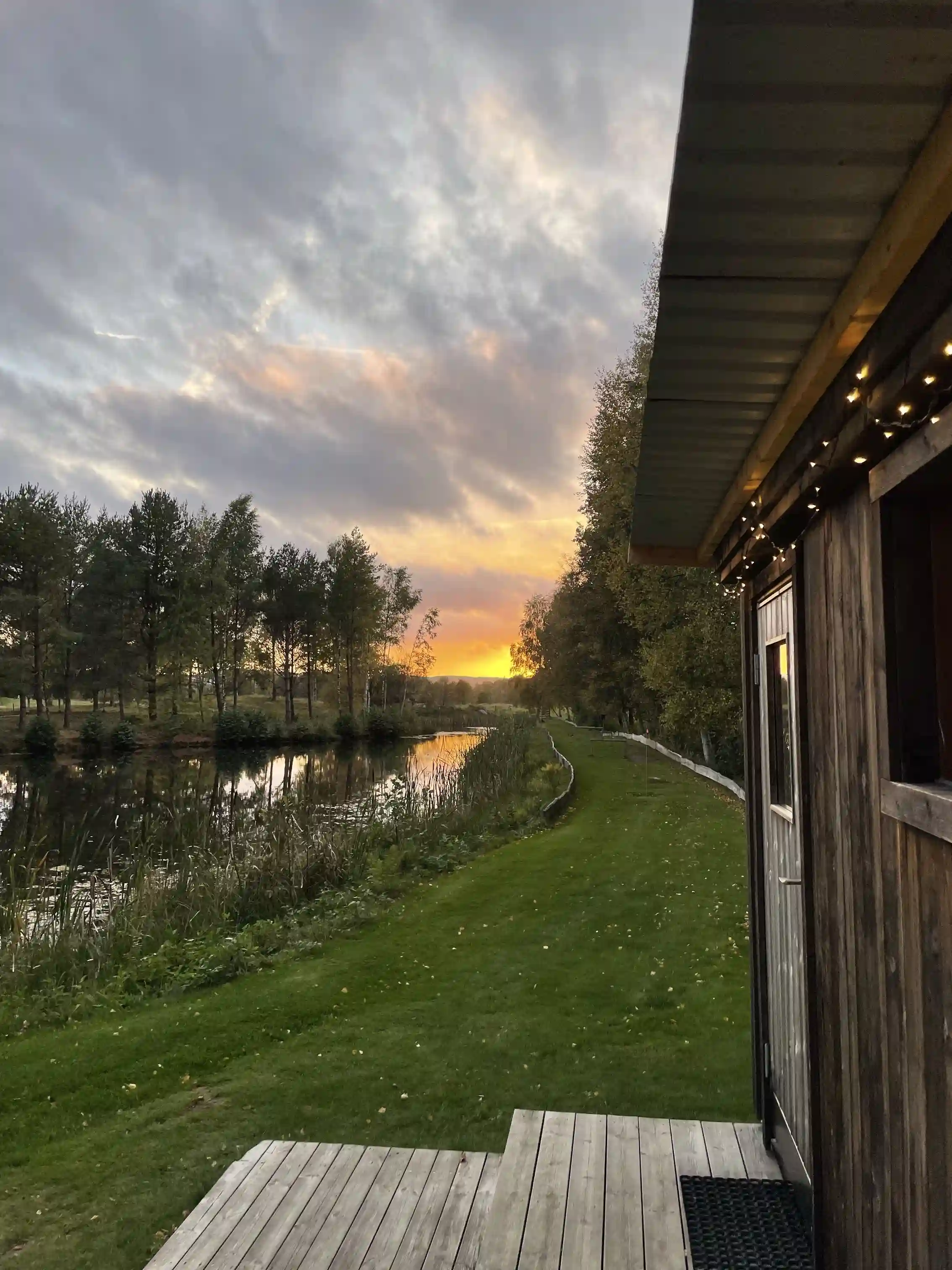 En stilla sjö omgiven av lummig grönska vid solnedgången på Våxtorps naturcamping. Spegelbilden av himlen och träden reflekteras i det lugna vattnet.