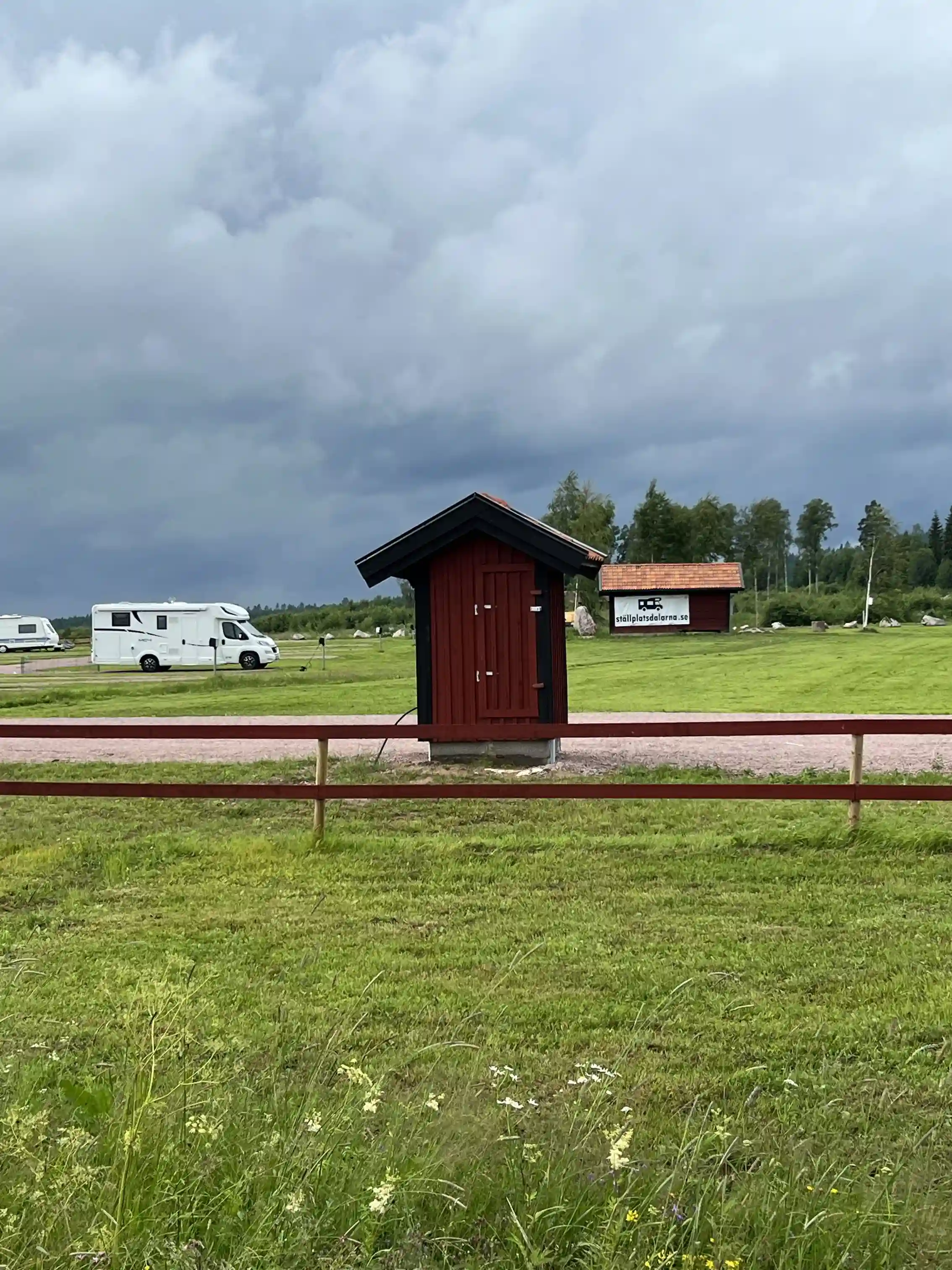 En vidsträckt grön äng i Dalarna, med en enkel byggnad och en grusväg i bakgrunden. Flera träpålar står uppställda i området, omgivna av riklig grönska. Perfekt landskap för en avkopplande utflykt eller campingupplevelse.