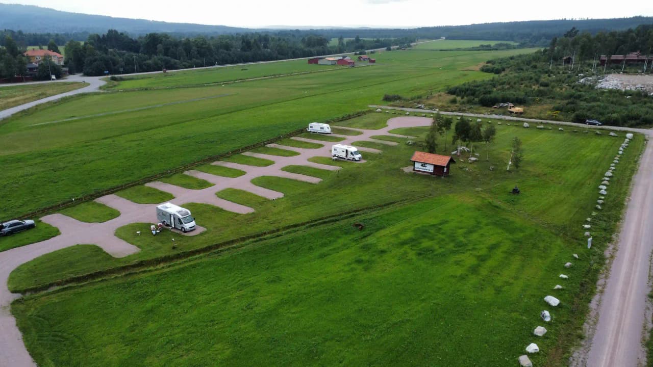 Flygbild av en ställplats i Dalarna omgiven av gröna fält och ängar. Parkeringsområdet är centralt beläget med lantlig omgivning, vilket inkluderar jordbruk och naturmarker.