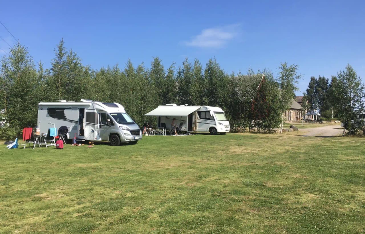 En husbil och en campingvagn är parkerade på en grön gräsmatta vid Bläsinge gård, omgiven av öppna fält i en campingmiljö.