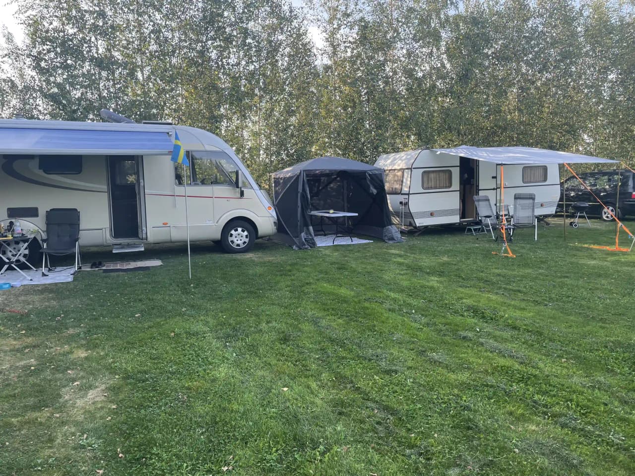 En lugn campingplats på Bläsinge gård med husbilar, en husvagn och tält uppställda på en grön gräsmatta. Träd skapar skuggiga områden och en bil är parkerad i närheten.