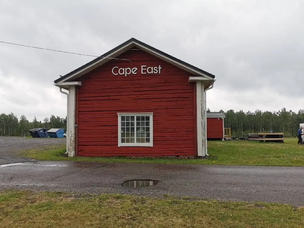 Bild som visar en lantlig miljö vid Cape East med en vidsträckt himmel och några moln. På marken syns en enkel byggnad, likt en stuga eller lada, med ett tak och fasad. Området omkring verkar öppet och lantligt.