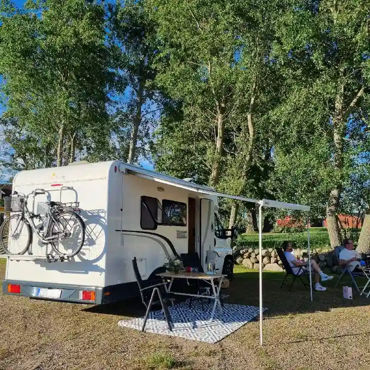 En husvagn står parkerad bredvid två cyklar vid en gårdsplan nära Björkängs vandrarhem. Cyklarna är placerade på grus med grönska i bakgrunden.