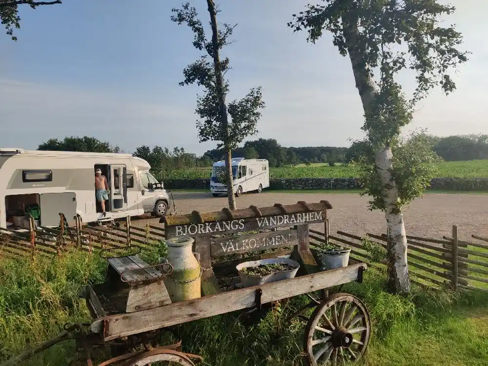 Bild av en traktordragen vagn på Björkängs vandrarhem. I bakgrunden syns gröna fält och en gård i lantlig miljö. Vagnen används troligen för aktiviteter eller transporter inom området.