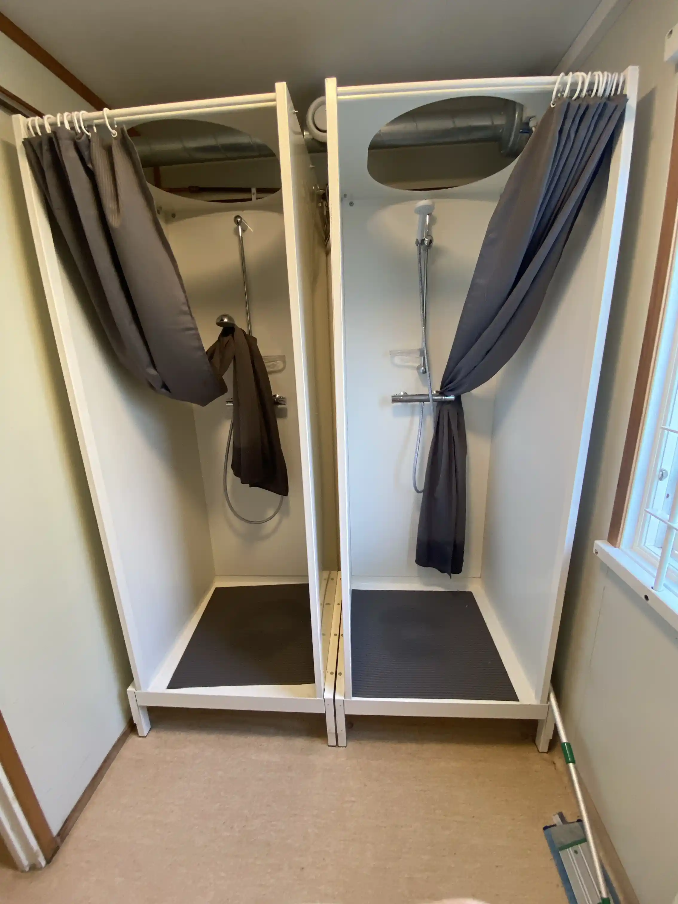 Ett enkelt och stilrent rum på Björkängs vandrarhem med grå färgschema, gardiner, en garderob och klädhängare. Rumsmiljön utstrålar renlighet och ordning.