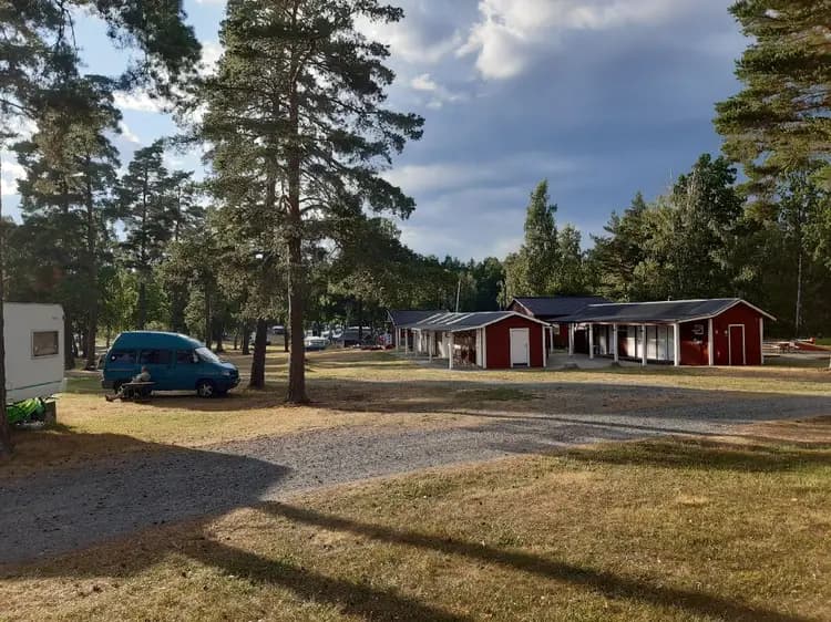 En somrig bild av Gudingebadet, där en liten stuga med tak syns bland träden. I förgrunden sträcker sig en grön gräsmatta, och i bakgrunden finns ett öppet landskap. Skuggor av träd ger en svalkande känsla åt platsen.