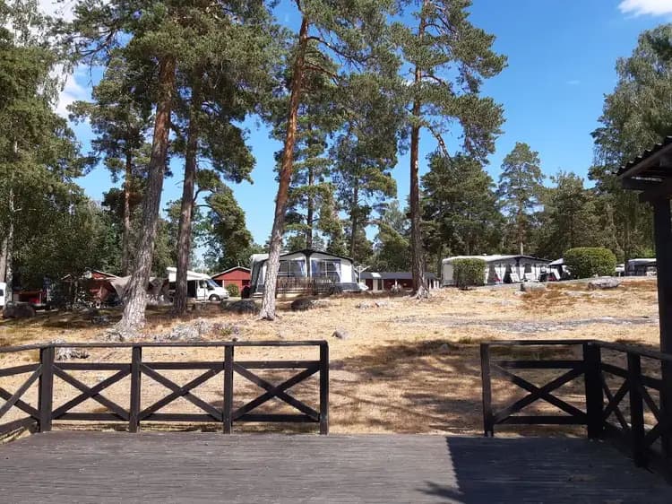 En lugn landskapsbild vid Gudingebadet med en stuga omgiven av träd och skugga, grusad uppfart, parkeringsområde och trädgårdsbyggnader som skapar en inbjudande rastplats.