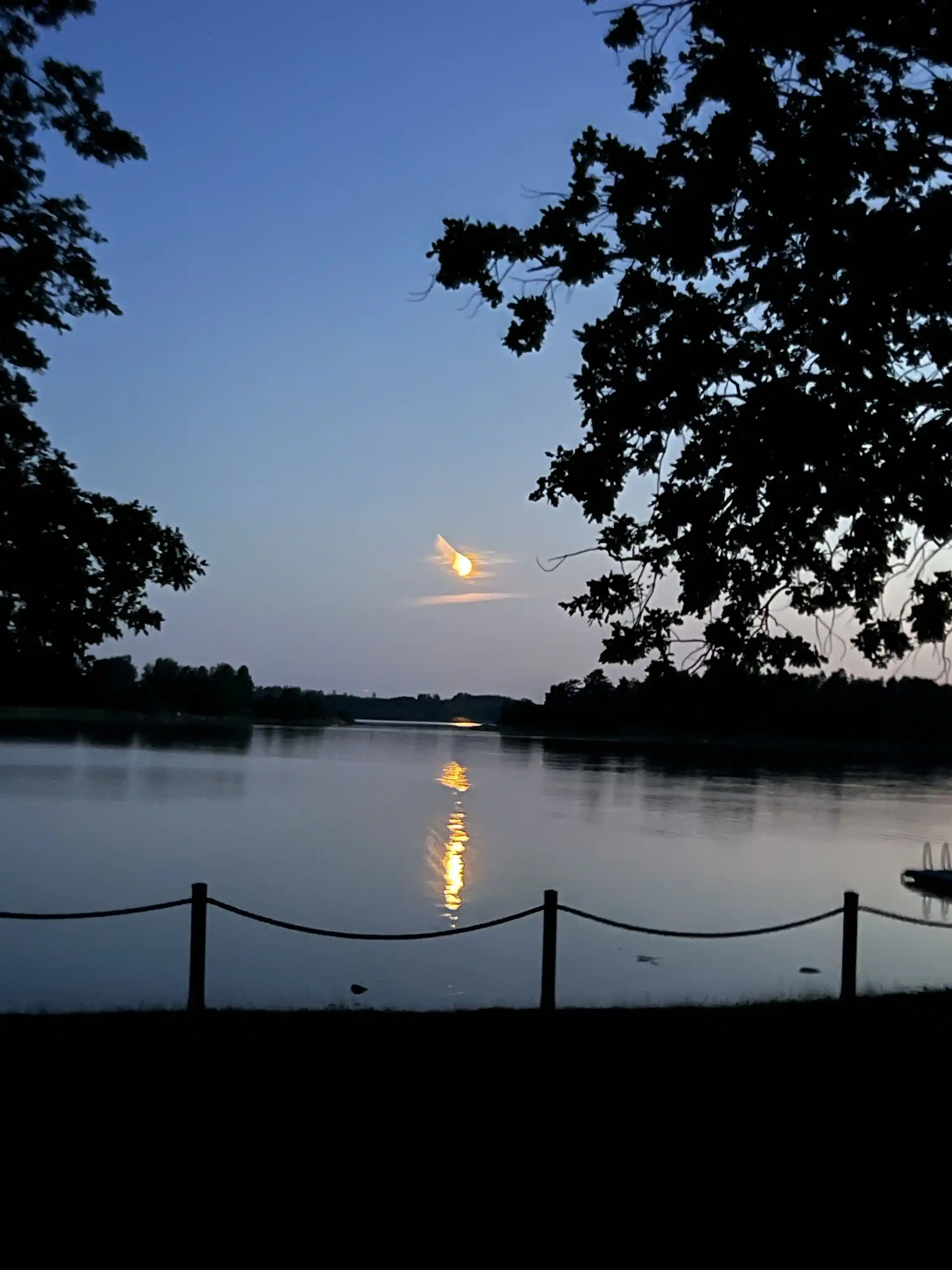 En solnedgång över Gudingebadet, där himlen speglas i det stilla vattnet, omgiven av träd och natur.