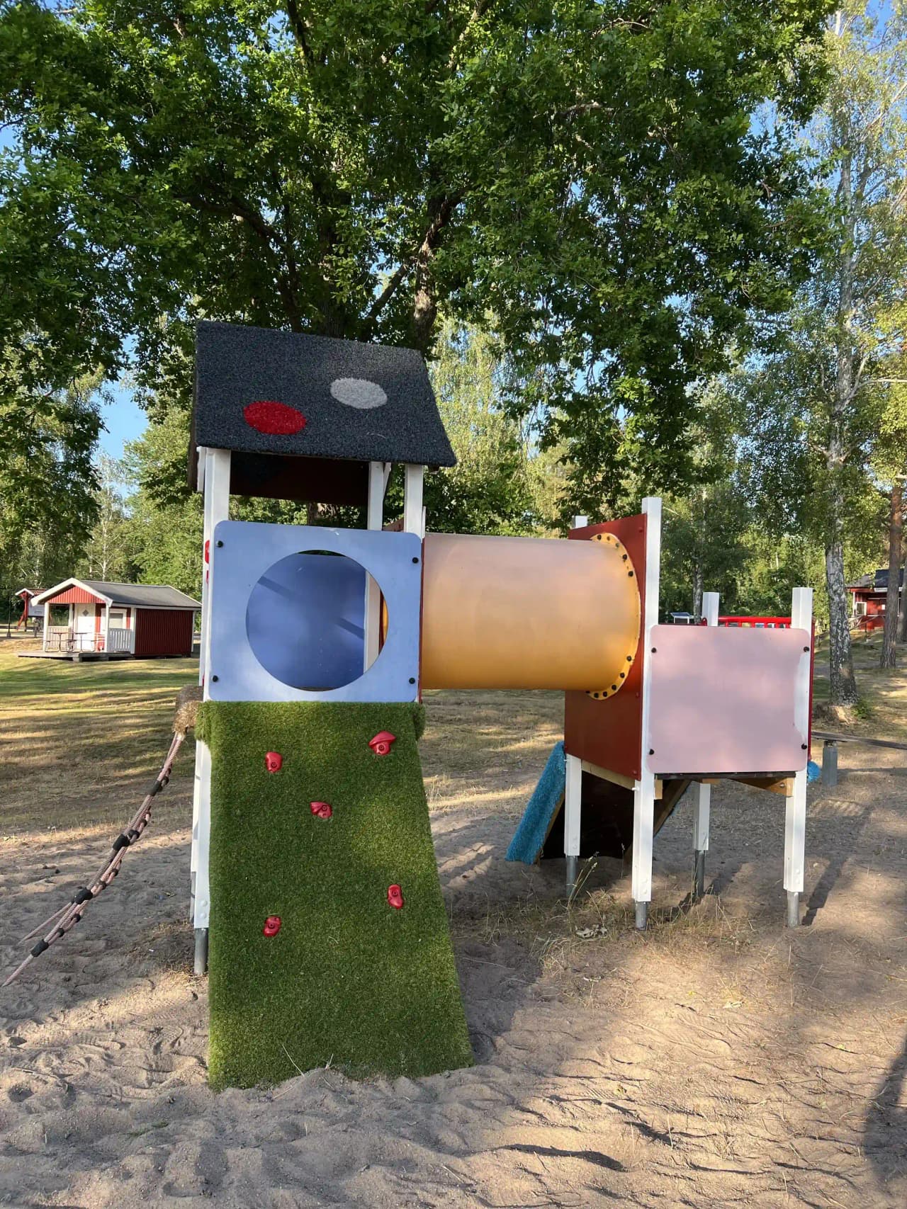 En lekpark vid Gudingebadet med rutschkana och andra lekredskap, omgiven av grönska och enstaka byggnader i bakgrunden.