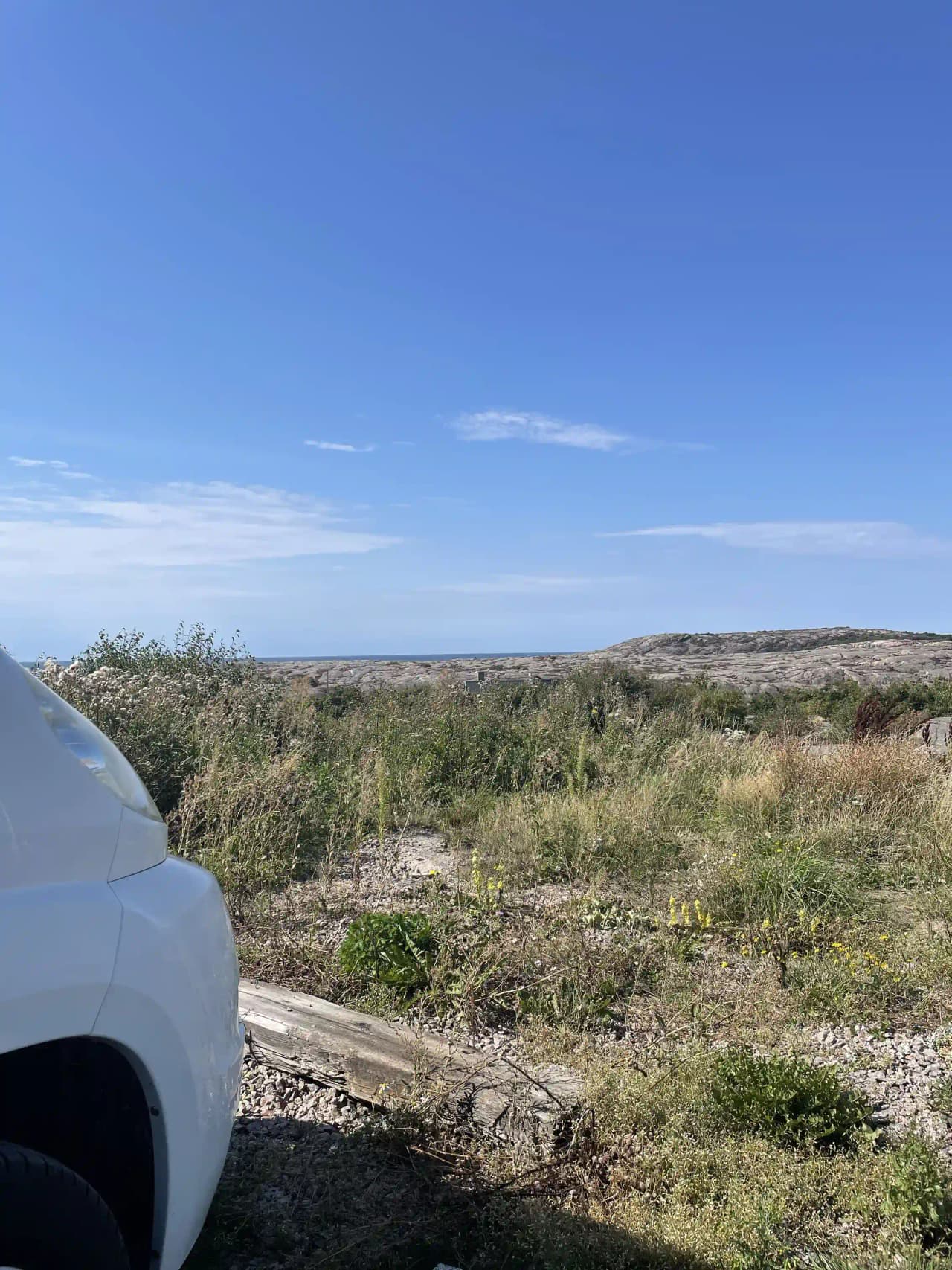 En SUV kör på en grusväg genom en solig naturlandskap vid Sotenäs Camping, med spegelbild i bilens sidobackspegel och omgivande vidsträckta steppemarker och buskage.