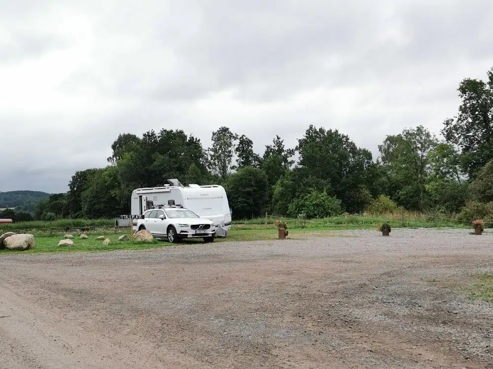 En vit skåpbil parkerad vid Gunnagård, omgiven av frodig grönska. Fordonet är i fokus och står på en grusväg med skog i bakgrunden.