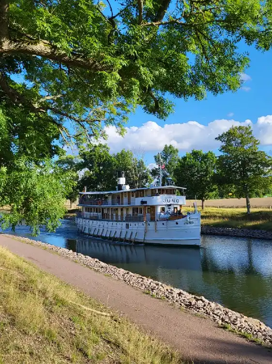 En båt flyter stilla på en klar kanal omgiven av grönska vid ett glasbruk, speglingar av båten syns i vattnet.