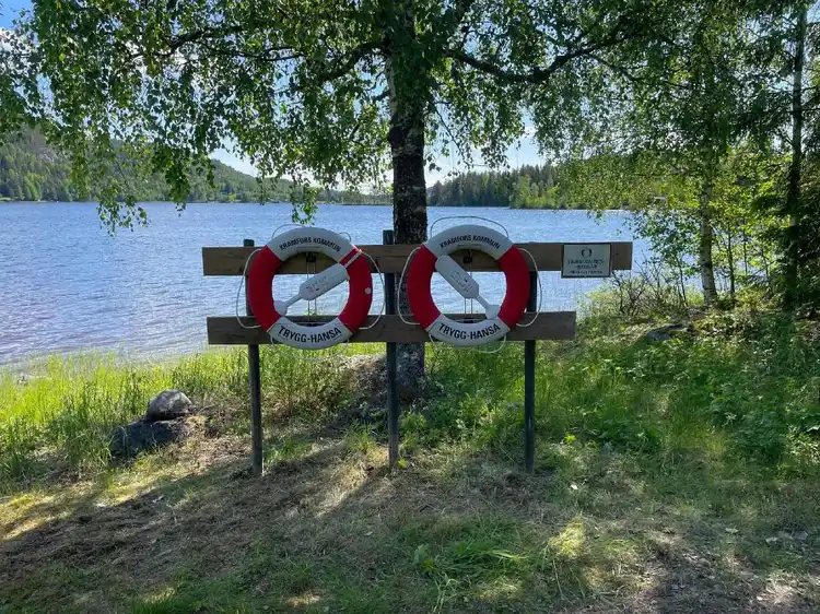 En bild av Valasjöns camping med en livboj i förgrunden och en sjö i bakgrunden. En skylt syns vid kanten av vattnet, omgiven av en naturskön miljö.