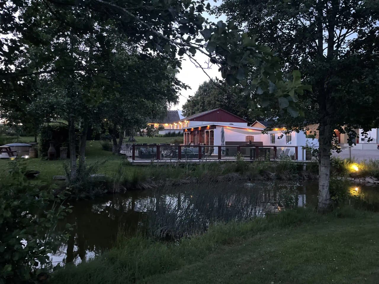 En naturskön bild föreställande Husargården, med en stuga omgiven av grönskande träd och buskar. En spegelblank damm speglas i omgivningarna, och skapar en rofylld atmosfär i naturmiljö.