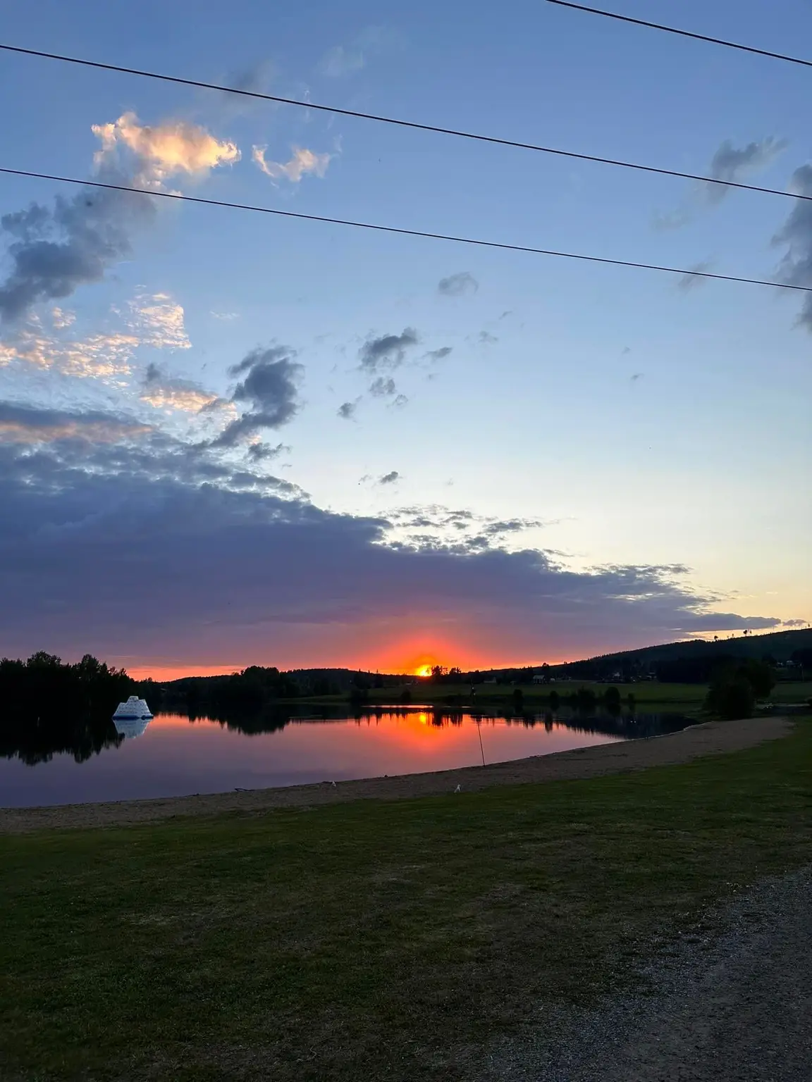 En solnedgång över Långsele Camping med en himmel i varma färger som speglas i det stilla vattnet, omgiven av natur och horisontlinje.