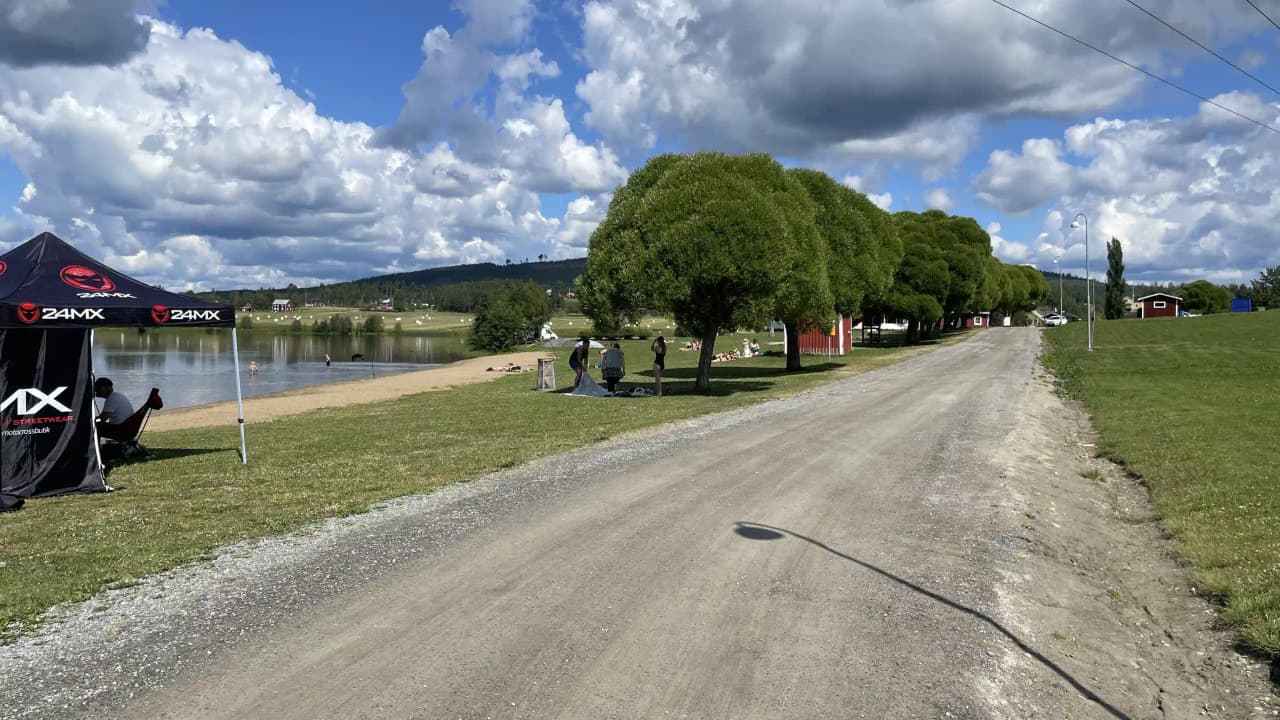 En campingplats vid en sjö med flera parkerade bilar, bland annat lyxbilar och sportbilar, omgivna av moln och grönskande landskap.