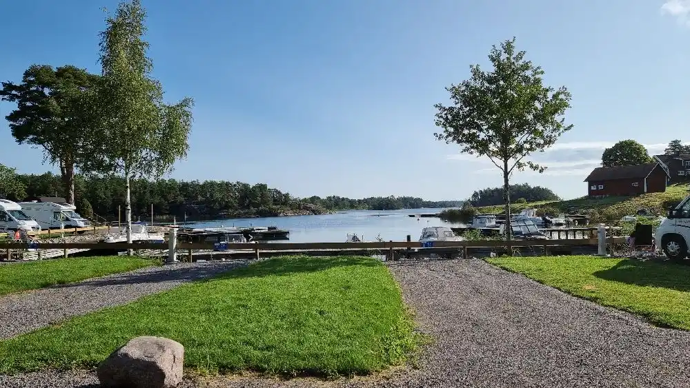 En solig dag vid Eriksviks hamn med båtar längs stranden och en lugn vattenspegel, omgiven av naturliga flodformationer.