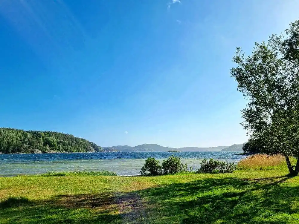Bild av Vindöns camping och marina: En somrig vy över kustlandskapet med blått vatten som speglas under en klar himmel. Grönskande vegetation omger strandkanten och ger platsen ett inbjudande och naturligt intryck.