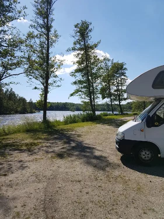 Bild av en fullsatt campingplats med olika fritidsfordon såsom husbilar och husvagnar parkerade på grusade ytor. Omgivningen är lummig med träd i bakgrunden och bilden ger en lugn utomhuskänsla, typisk för en campingmiljö.