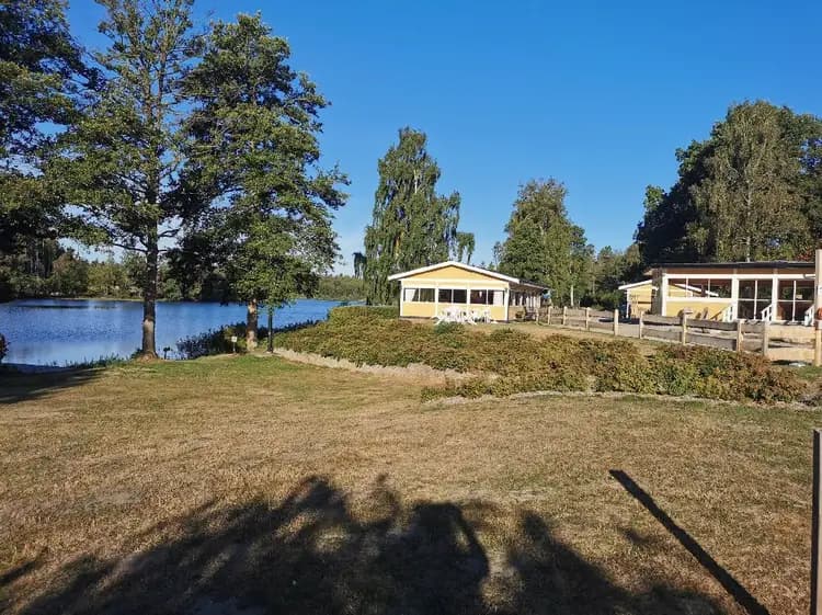 En grön camping med träd och gräs i förgrunden, små stugor ligger utspridda på området. I bakgrunden syns en sjö som omges av skog.