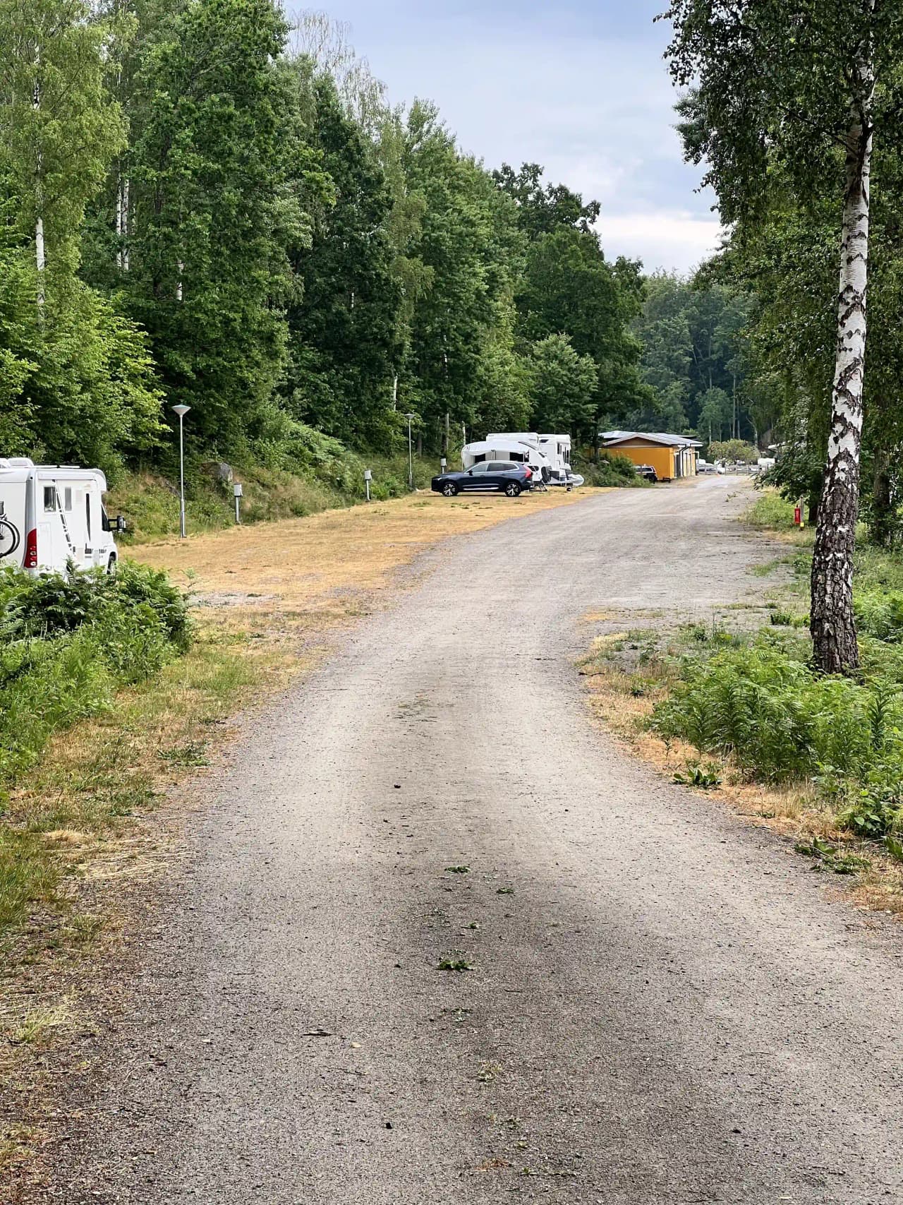 En grusbelagd campingplats vid Ljuvadal med träd i bakgrunden, där flera husbilar och husvagnar är parkerade längs en asfalterad väg.