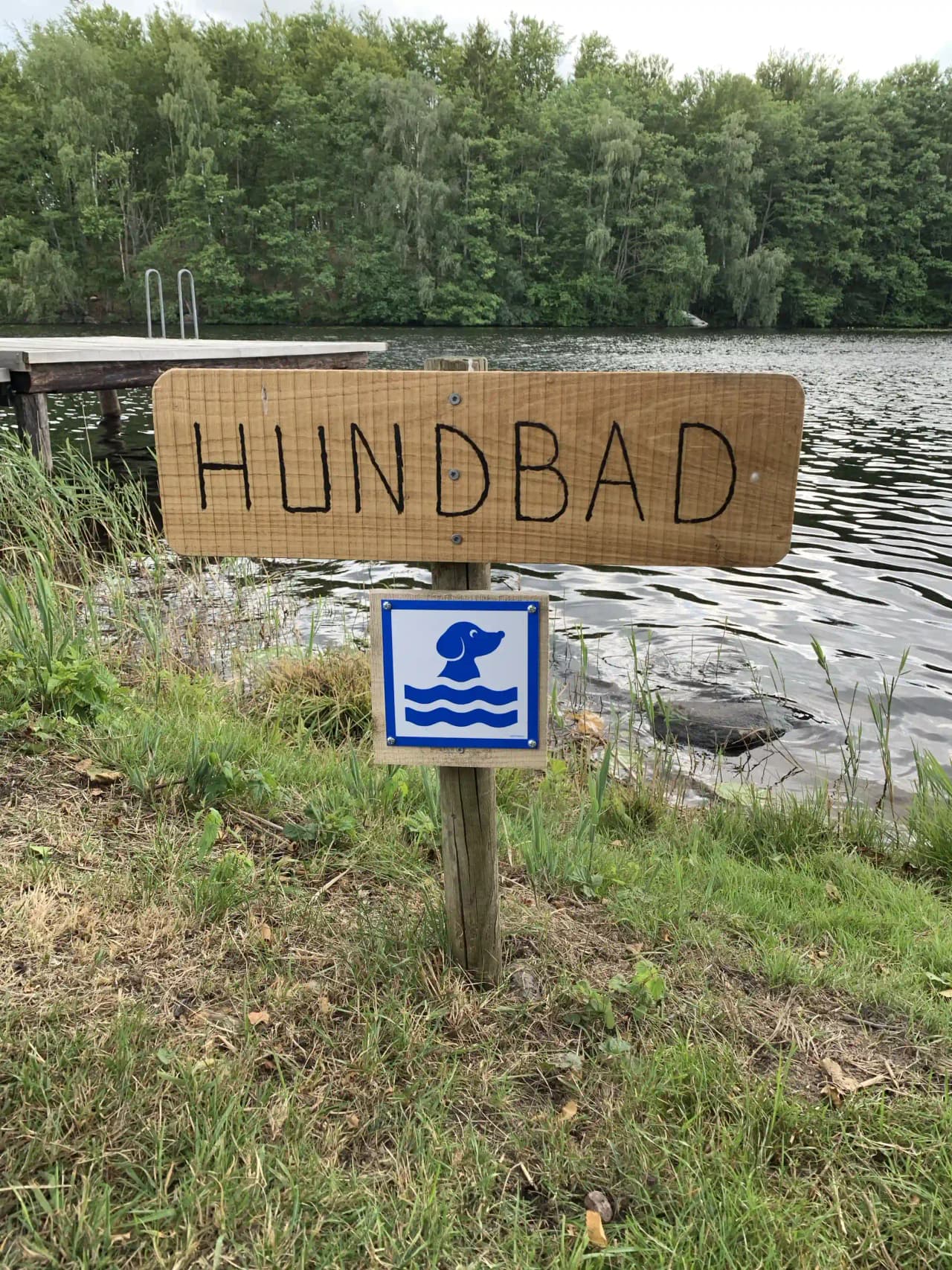 Vy över Ljuvadal Camping & Ställplats med en naturskön sjö i bakgrunden. Vegetation omger vattenområdet och i förgrunden syns en skylt som markerar platsen.