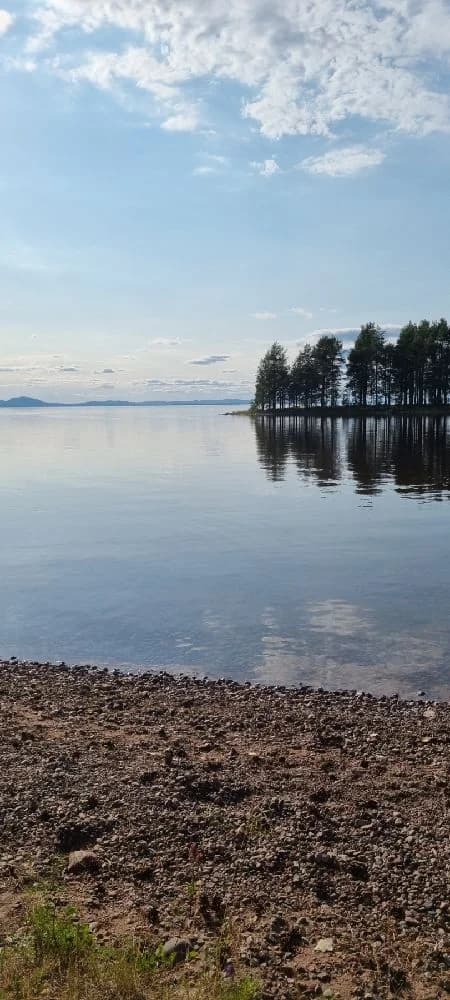 En klarblå himmel speglas i det lugna vattnet vid Tällbergs camping, med moln som svävar vid horisonten.
