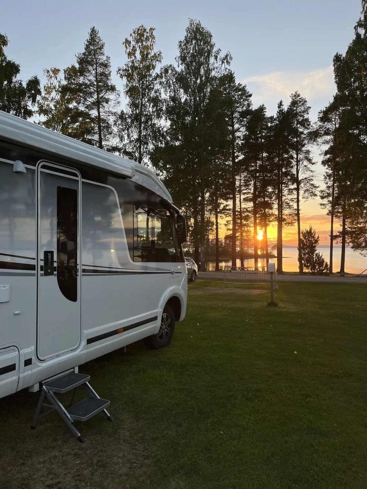 Campingplats vid sjön på kvällen med flera husvagnar och campervans uppställda på en grön gräsmatta. Solnedgångens ljus skimrar i bakgrunden, vilket skapar en rofylld och avkopplande atmosfär.