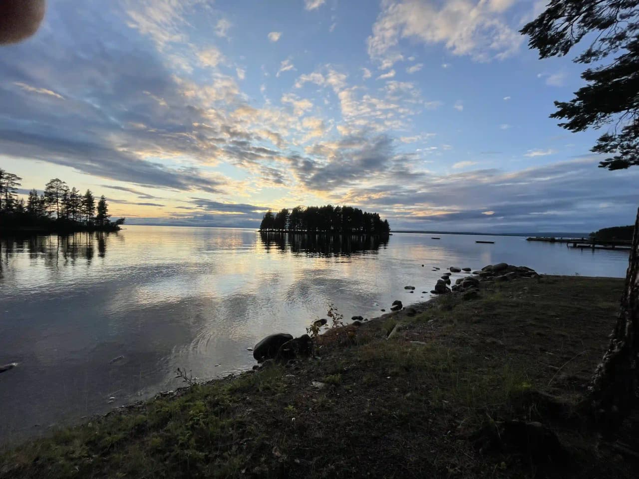 En vy över sjön med spegelblankt vatten och en klar blå himmel, omgiven av moln. Naturen sträcker sig längs horisonten, vilket ger en fridfull atmosfär vid Tällbergs camping.