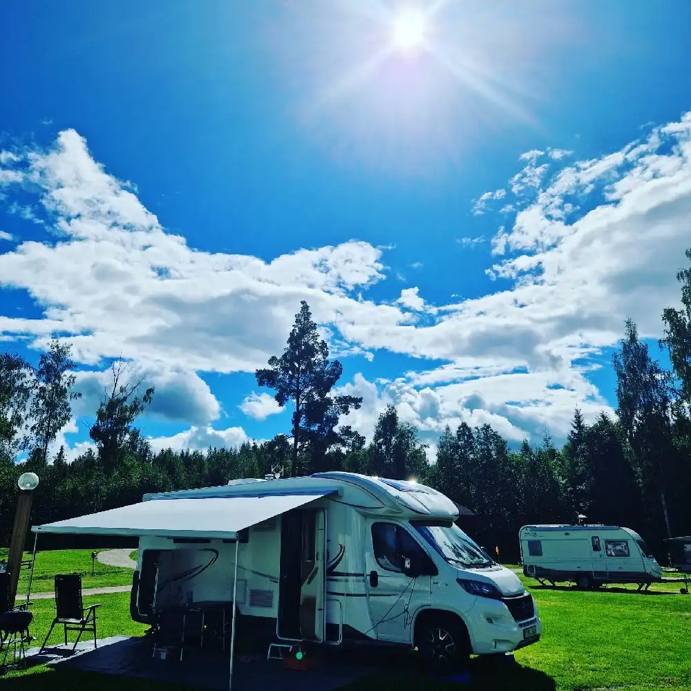 Bild av en husbil och husvagn uppställda på Tällbergs camping omgivna av grönska och campingutrustning i en naturnära miljö.
