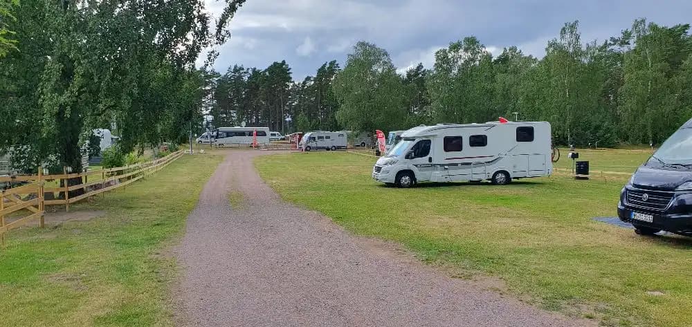 En campingplats med flera husbilar och husvagnar parkerade på en grusbelagd yta, omgivna av grönskande träd och natur.