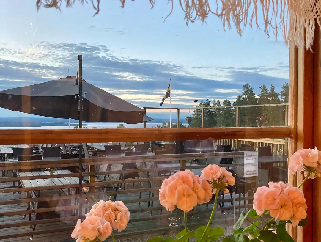 En balkong med träyta som tillhör Åsengården, med stolar och ett utomhusbord i skugga. Blomkrukor och husväxter bidrar till den hemtrevliga atmosfären, vilket ger en känsla av ett eco-hotell med restaurang.