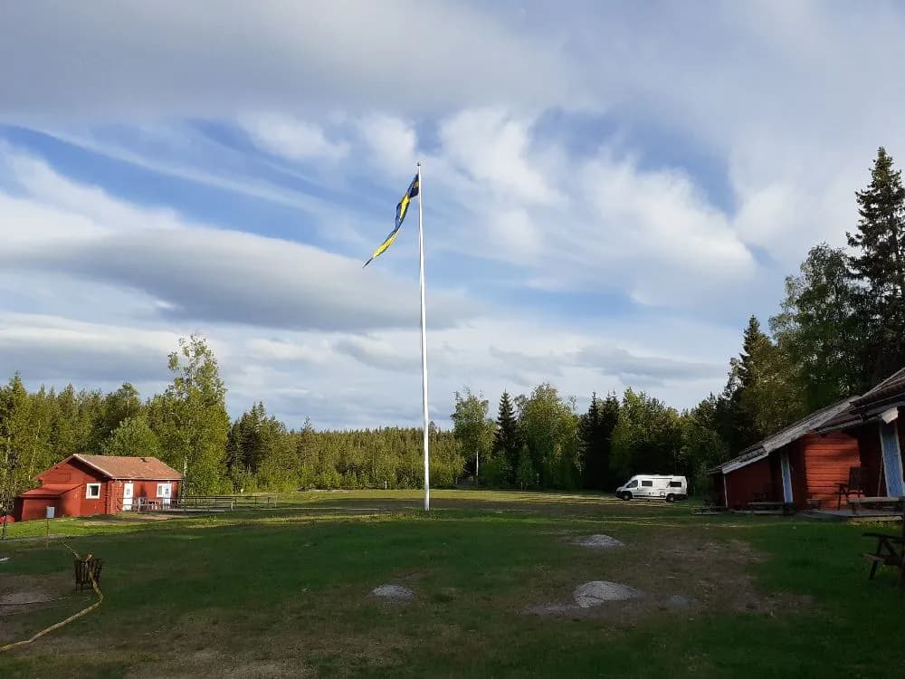 En solig dag vid Åsengården med en flaggstång mot en blå himmel med cumulusmoln, omgivna av en grönskande gräsmatta och barrträd i bakgrunden.