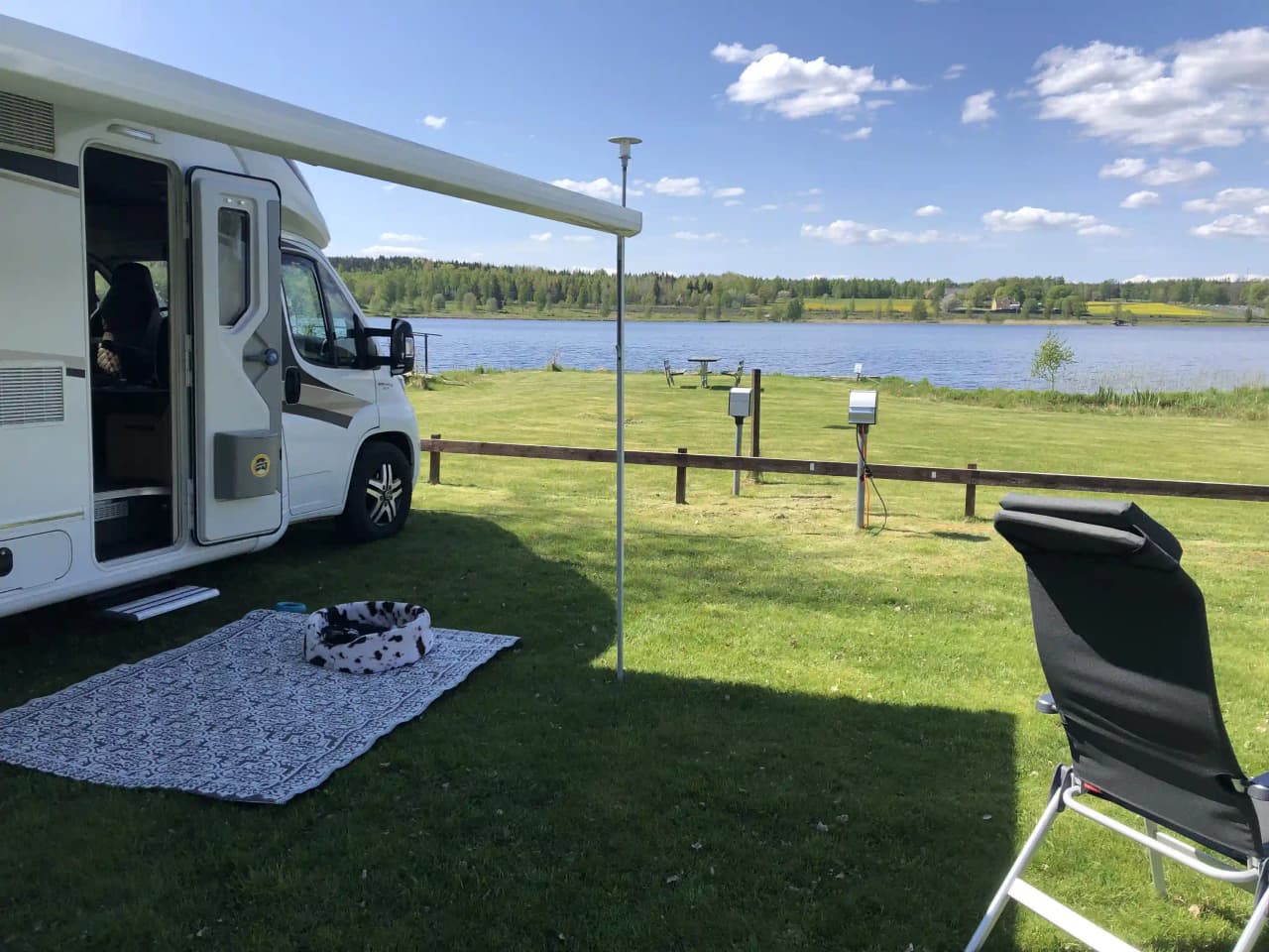 En campingpark med grön gräsmatta där flera husvagnar och fritidsfordon står parkerade i skuggan. Det finns utemöbler uppställda på gräsmattan, vilket skapar en inbjudande och avslappnad atmosfär.