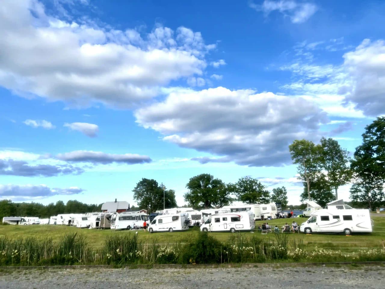 En bild av Upperuds Gästhamn visar en naturskön plats med molntäckta himlar, stora ängar och en parkeringsplats där flera husvagnar och en lätt lastbil är uppställda.