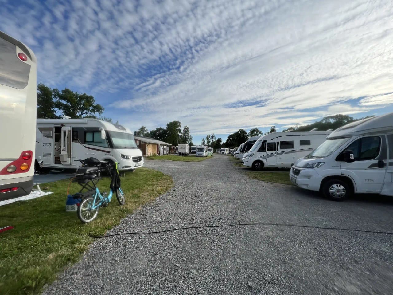 Bild på Upperuds Gästhamn med flera husbilar och campingfordon parkerade på en grusplan. Området är omgiven av grönska och himlen är klarblå.