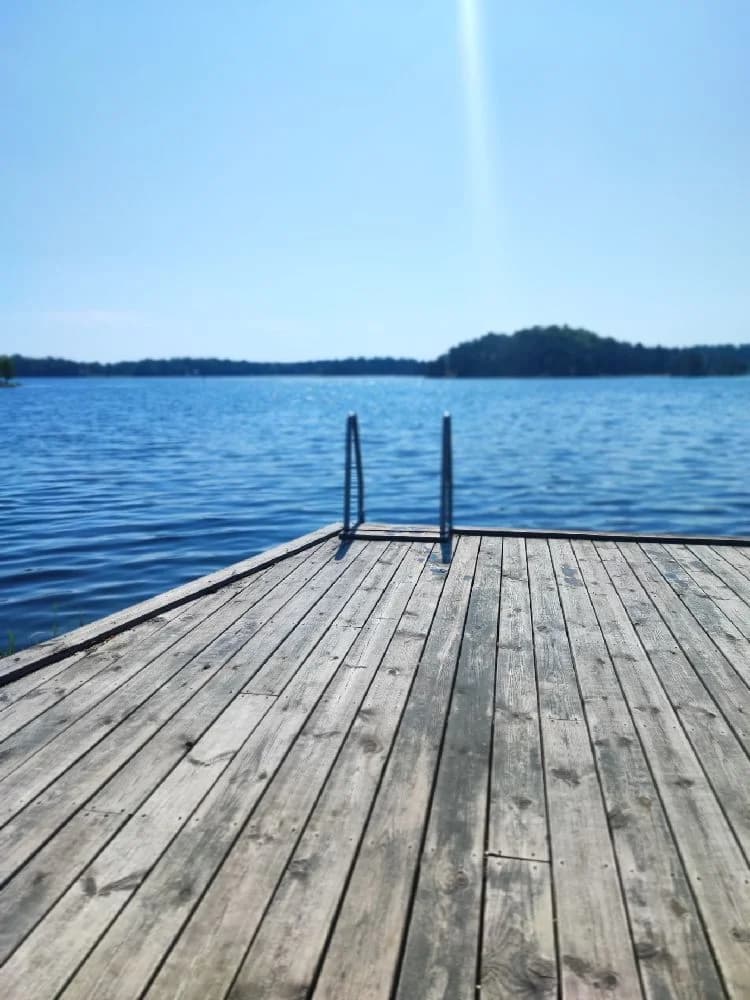 En träbrygga sträcker sig ut över ett spegelblankt blått vatten vid Upperuds Gästhamn, omgiven av en naturskön omgivning. Bryggan består av välunderhållna träplankor och bidrar till den idylliska stämningen vid vattnet.