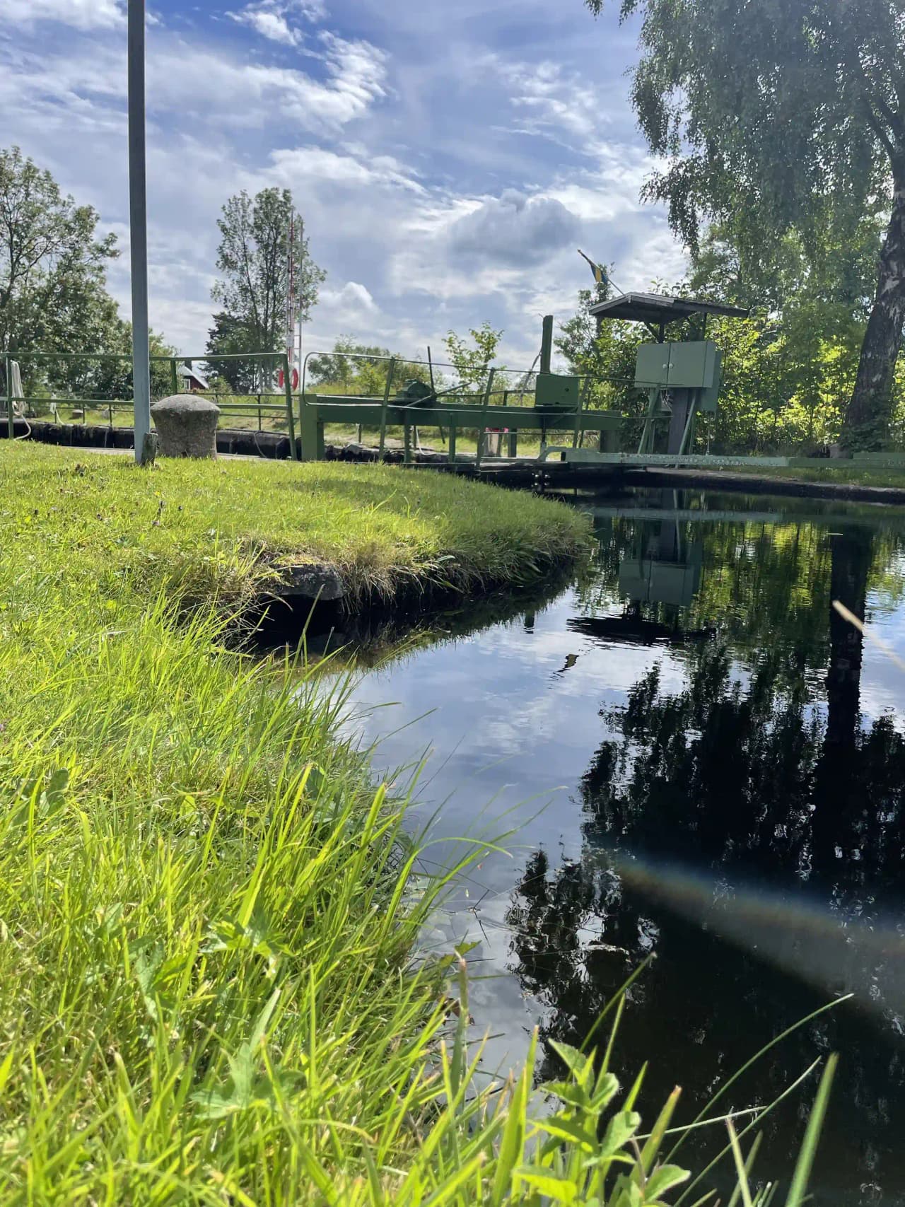 Bild av Upperuds Gästhamn: en lugn vattenkanal omgiven av grönskande växter och gräs, med spegelklara reflektioner i vattnet och en välansad trädgård i närheten. 