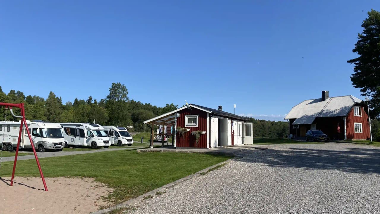 Bild av Upperuds Gästhamn: En grönskande campingplats med husvagnar och husbilar parkerade på ett gräsfält. En väg leder igenom området, och det finns gott om utrymme för rekreation och avkoppling.