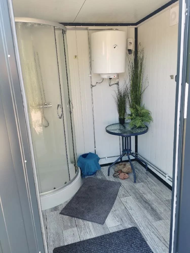 En modern och ren badrumsmiljö med vita kakelväggar och ett fristående toalettstol med stängt lock. Till höger finns en dusch med aluminiumram och glasdörr. Fokus ligger på enkelhet och funktionalitet.